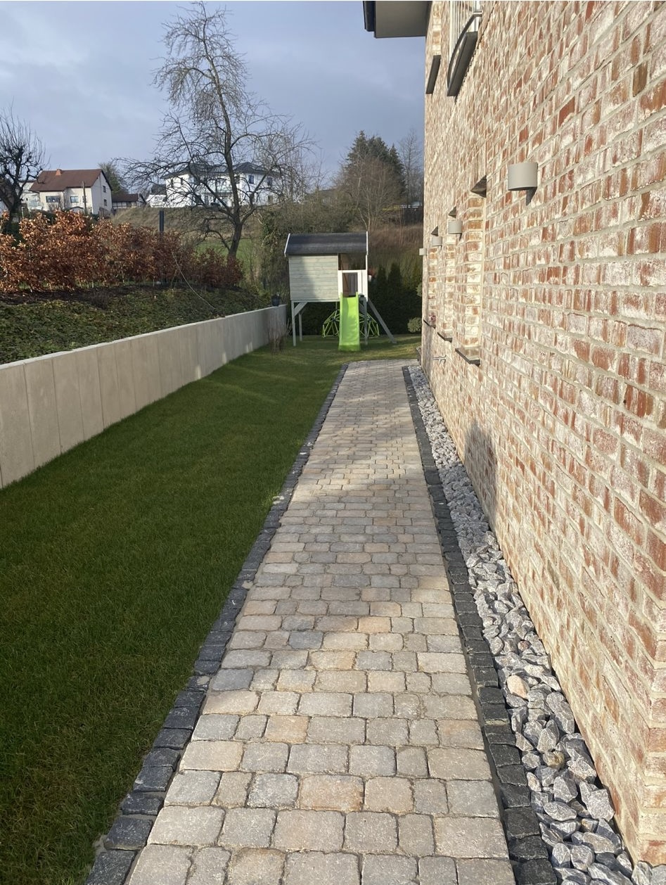 Neu verlegter Natursteinpflasterweg mit Randsteinen und Kiesstreifen entlang einer Klinkerfassade